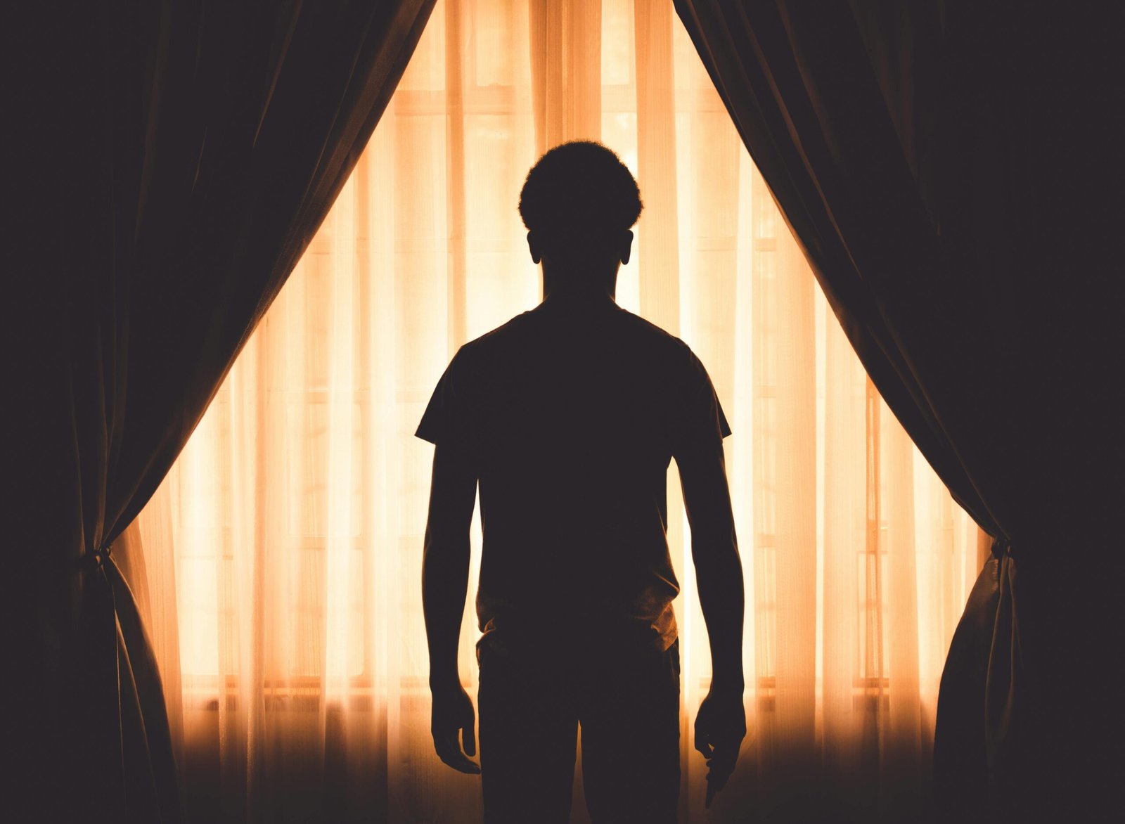 A silhouette of a man standing by a window with curtains, creating a dramatic contrast and mood.